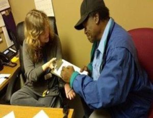 Facilitator and consumer in the PACE office. The faciliitator is holding a clipboard for the consumer as he completes paperwork.  